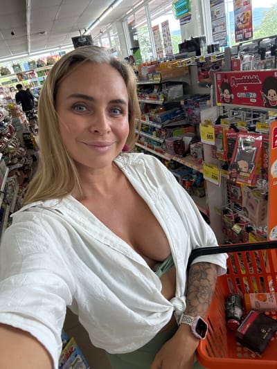 In Thap Sakae, Thailand, a fair-skinned Caucasian woman with blonde hair and grey streaks is taking a selfie in a convenience store. She is wearing an unbuttoned white linen shirt, revealing a green bralette. She has a floral tattoo on her left arm and is wearing a smartwatch. She is holding the handle of an orange shopping basket that contains various items, including a small dark wallet, a tube of sunscreen, and a can. In the background, shelves are stocked with merchandise. The store appears well-lit with fluorescent lighting and sunlight streaming in from windows. Another person, a man in a dark t-shirt and patterned shorts, is visible in the background, looking at the shelves. The environment is typical of a retail establishment, with rows of products and aisles.