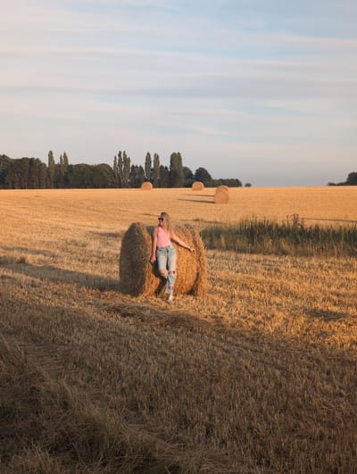 In Alvimare, France, a young Caucasian woman with blonde hair stands leaning against a round bale of hay in a field of dry, cut wheat. She is wearing a pink tank top and ripped blue jeans. Her sunglasses are on her head. The field is filled with rows of hay bales, some closer and more detailed, others further in the background. A line of trees forms the horizon behind the field, under a pale blue sky with wispy clouds. The sun is low, casting long shadows and bathing the scene in a warm, golden light.