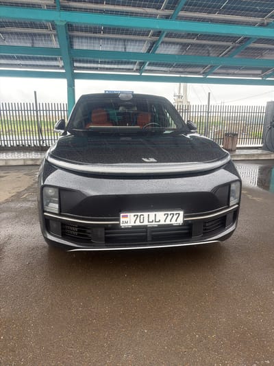 The image shows the front of a black car with an Armenian license plate "70 LL 777". The vehicle is parked on a wet surface. The background includes a fence and a structure with a blue metal frame and translucent panels, possibly a carport or covered parking area. The location is potentially Tsovagyugh, Armenia, based on the license plate origin. No people are visible.