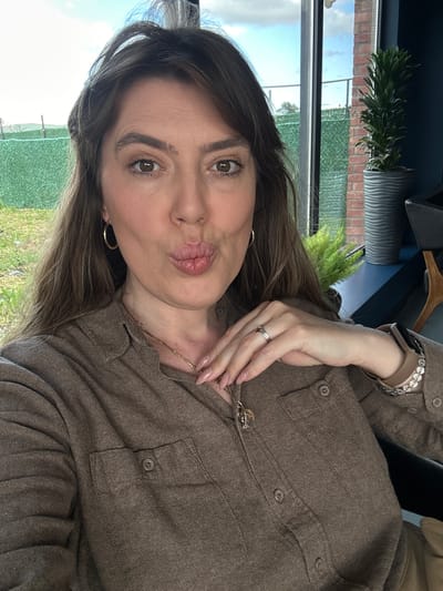 A woman is posing indoors for a selfie. She is looking at the camera with puckered lips. She has long brown hair, gold hoop earrings, a gold chain necklace, and is wearing a brown button-down shirt. She also wears a tan watch and a ring on her left hand. Outside the window behind her, there is grass, fencing, and trees. Brick buildings are visible in the background. The location in Marneuli, Georgia cannot be confirmed from the image.