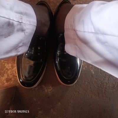 This media file shows a close-up, overhead view of a person's feet wearing black, glossy loafers. The loafers have a light-colored sole. The person is wearing light-colored pants, possibly white or off-white, which appear slightly rumpled. The background is a rough, textured surface, likely a floor, with shades of brown and reddish-brown. The lighting suggests it could be daytime, with natural light reflecting off the shoes. The setting appears to be indoors, given the texture of the floor and the way the light falls. There is no discernible activity taking place other than the person being stationary. The image includes a watermark in the bottom left corner that reads "LEXDEN DIGITALS". The overall impression is a candid shot focusing on footwear and attire, with a simple, everyday setting. No specific emotions are conveyed. The location context provided is Edu Town, Nigeria.