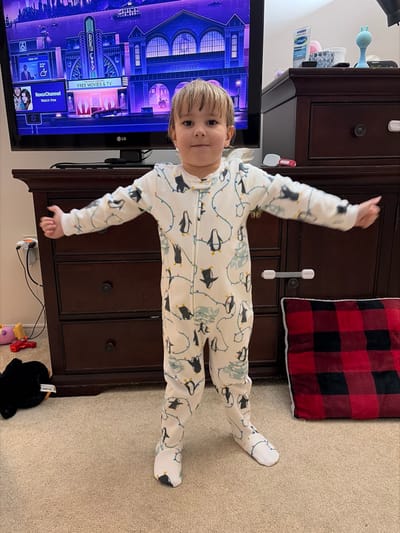 An image taken indoors, within a residential setting in Woodstock, Canada, depicts a young child standing centrally on a light brown carpeted floor, facing the camera directly. The child, a toddler with light brown hair and dark eyes, wears a white, footed sleepsuit patterned with black-and-white penguins and colorful string lights. The child has both arms outstretched horizontally to the sides, palms slightly upward, and a slight smile.

Behind the child, a black flat-screen television sits on a dark brown wooden dresser or media console. The TV displays a digital cityscape scene, labeled "Roku City," with purple and blue tones, along with "Roku Channel" branding and "FREE MOVIES & TV" text. A weather widget shows "22°." To the right of the television, on the dresser surface, are a white tube of Cetaphil cream, a grey textured container, and a light blue silicone facial scrubber. A white child safety latch is affixed to the top right drawer of the dresser.

On the carpeted floor to the child's left, a black plush toy, a pink toy, and a red toy with a yellow tip are visible, along with white electrical cords leading to a wall outlet. To the child's right, a red and black buffalo plaid cushion or pillow with white trim rests on the floor.