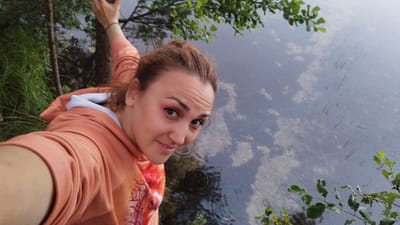 A high-angle medium shot shows a young woman with reddish-brown hair in a loose bun, facing the camera and smiling. She is wearing a coral-colored hooded sweatshirt with a white lining at the neck. The woman is outdoors, with her left arm extended as if taking a selfie. To her right, a body of water, likely a lake or river, fills the majority of the frame, with reflections of the sky and surrounding vegetation visible on its surface. To the left of the woman, tall green grass and a thin tree trunk are visible. Green leaves from a tree branch hang into the upper right corner of the frame, partially obscuring the water. The overall scene suggests a natural, outdoor setting.