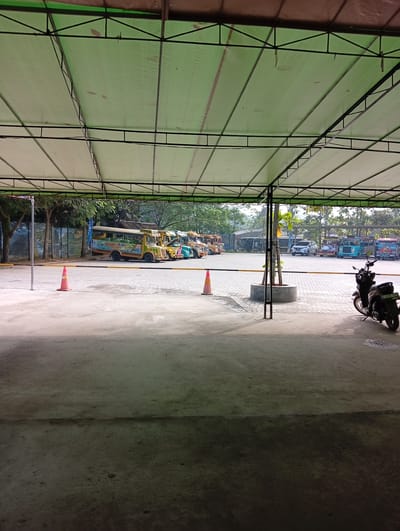 The image shows a covered parking area in Parung Panjang, Indonesia. Several brightly colored "Kereta Baliabahari" buses are parked along the far left side. Two orange traffic cones are placed near the center. A black motorcycle is parked on the right. In the background, other vehicles, including cars and additional buses, are visible. The area is paved with concrete and bricks. The structure overhead is a green metal frame covered with a green tarp.