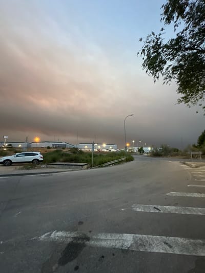 The image captures an industrial area on the outskirts of Cabanillas del Campo, Spain, under a hazy sky tinged with orange and pink hues, suggesting smoke or dust. A white SUV is parked on the left side of the road, beside a patch of overgrown vegetation. In the background, a row of modern industrial buildings with white facades and dark roofs stretches across the horizon, illuminated by streetlights and possibly distant fires. The foreground shows a paved road with white pedestrian crossing markings. A large tree with dark green foliage is visible in the upper right corner of the frame, casting shadows. The overall atmosphere is subdued, with muted colors and a sense of twilight.