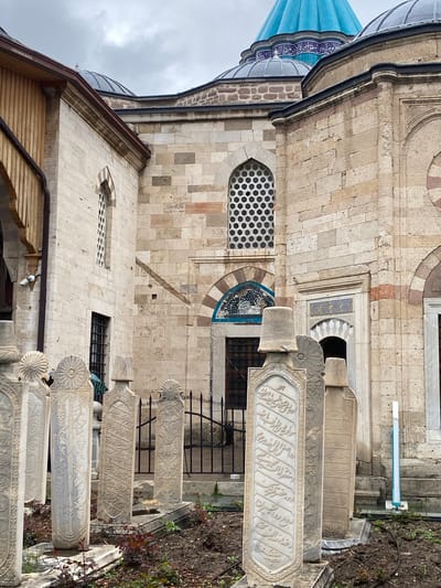The image depicts a section of the Mevlana Museum in Karatay, Turkey. The architecture features tan-colored stone walls with arched windows and geometric patterns. A portion of a blue dome is visible on the left side of the frame. Adjacent to the building, there is a row of intricately carved stone tombstones with inscriptions. Some greenery is present on the right side of the frame. The overall scene appears to be a historical or religious site.