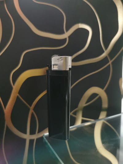The image shows a black lighter with a silver ignition mechanism that has the letters "AGS" embossed on it. The lighter is positioned vertically on a clear glass surface, and a black background with gold, swirling patterns is visible behind it. The scene is indoors, and the lighting suggests it might be evening or a dimly lit room. The origin of the image is unknown, and there is no indication of its location in Bourg-lès-Valence, France.