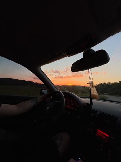 The image depicts the interior of a car with a person's arm on the steering wheel, driving towards a sunset. The sky is filled with orange and pink clouds, with the sun just above the horizon. The dashboard lights are on, and a car air freshener hangs from the rearview mirror. The scene appears to be a rural road, possibly in Hrabovets, Ukraine, given the rolling hills and vegetation visible outside the car windows. The overall mood is peaceful and contemplative, with the warm sunset colors creating a serene atmosphere.