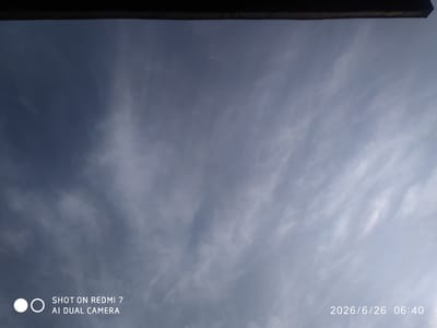 This media file depicts a sky with wispy, thin clouds, suggesting a partly cloudy or overcast day. The sky is a pale blue, transitioning to a lighter shade near the horizon, implying it is either early morning or late afternoon. A dark, horizontal object is visible at the very top of the frame, possibly the edge of a building or a rooftop.
In the bottom left corner of the image, there is a circular logo with two solid dots and a circular outline, typical of camera branding. Adjacent to this logo, text reads "SHOT ON REDMI 7 AI DUAL CAMERA". In the bottom right corner, a timestamp is visible: "2026/6/26 06:40".
The scene appears to be an outdoor setting, likely captured from a low angle looking upwards. The clouds do not indicate any severe weather. The timestamp suggests the image was taken at 6:40 AM on June 26, 2026. The location context provided is Khalkabad, Uzbekistan. No people or specific objects are discernible in the sky itself, apart from the clouds. The overall mood is calm and observational, focusing on the natural sky.