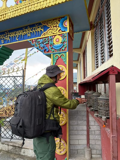 A male figure is depicted in a three-quarter profile view from the left, standing before a series of prayer wheels in Ghandruk, Nepal. He wears an olive-green hooded jacket, dark green trousers, and a dark grey bucket hat, and carries a large black backpack. His right hand is placed on the leftmost of four dark, cylindrical prayer wheels, which are intricately carved with script and housed under a red, pitched canopy structure attached to a light yellow building. The building features multiple brown louvered windows. To the left of the figure is a vibrantly decorated archway entrance, adorned with gold, blue, red, and green traditional designs, including golden Tibetan script on a central blue band. Beyond the archway, numerous colorful prayer flags are strung against a cloudy sky, with glimpses of distant buildings. The immediate ground consists of light grey stone steps and a paved surface.