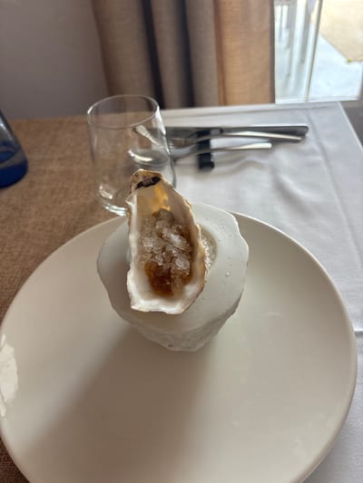 A close-up, eye-level shot features a single oyster served in its shell, resting on a white, abstract pedestal, which in turn sits on a large white plate. The oyster is filled with ice shards and a dark, viscous liquid. The plate is positioned on a white tablecloth, with a textured brown tablecloth visible in the background to the left. To the left of the plate, a clear glass is placed on the brown tablecloth. Behind the glass, a set of cutlery rests on the white tablecloth. A window with beige curtains is visible in the upper right corner of the frame. The overall lighting suggests an indoor setting. This scene is likely from a restaurant in Carboneras, Spain, given the presentation and typical dining setup. There are no people visible in the image.
