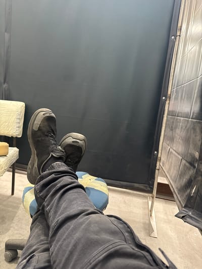The image shows a person sitting in a chair. The person is wearing gray pants and black shoes. The chair has a blue cushion. The background is a black curtain or wall with a metal frame. The location is unknown, and there is no information to suggest it is in Račice, Czechia.