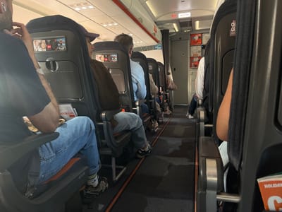 The image shows the interior of an airplane cabin. The seats are occupied by passengers. The aisle is visible, and there is an exit sign. The seats have headrests with advertisements. The location is likely Lyon–Saint-Exupéry Airport in Colombier-Saugnieu, France.