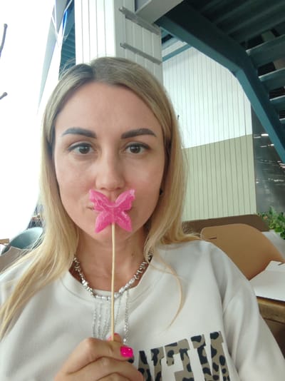 A woman with blonde hair and dark eyebrows is holding a pink butterfly-shaped lollipop in front of her mouth. She is wearing a white sweatshirt and a silver necklace. The background appears to be an indoor setting with a white and beige striped wall and a blue staircase. The image is a selfie taken from the chest up.