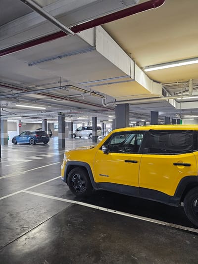The image depicts a bright yellow Jeep Renegade parked in a multi-story car park. The car is positioned at an angle, with its driver's side visible. The interior of the car is partially obscured by dark tinted windows, but the steering wheel and dashboard are discernible. The car park itself is characterized by a concrete floor with white painted parking lines, concrete pillars, and extensive overhead ductwork and piping, suggesting an industrial or functional environment. Fluorescent lighting illuminates the scene, casting long shadows. Further down the aisle, two other cars are visible: a dark blue hatchback and a lighter colored car. A red pipe runs along the left side of the image, contrasting with the neutral tones of the car park structure. The overall impression is that of a typical indoor parking facility, possibly in a commercial or residential building. No people are visible in the image. The time of day is indeterminate due to the indoor setting, and there is no indication of weather conditions. The location context provided is Cuarte De Huerva, Spain.