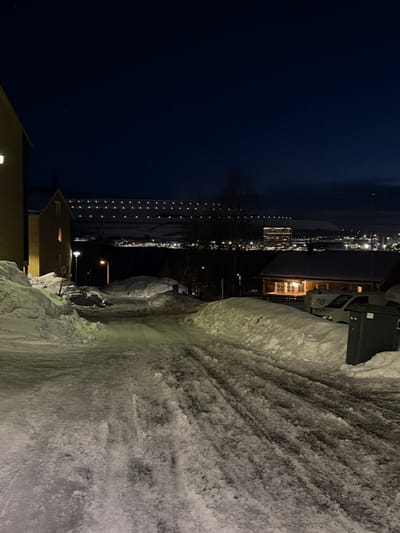 The image depicts a snow-covered residential street in Kiruna, Sweden, at night. The foreground road is packed with snow and ice, exhibiting tire tracks, and slopes downwards. Substantial snow banks line both sides of the roadway. On the left, a light-colored building displays an illuminated window, with a streetlamp casting light onto the immediate area. Further back on the left, another building is partially visible. On the right, a darker-colored house features an illuminated porch or window area. Two white panel vans are parked on the right, partially obscured by snow, adjacent to a dark grey refuse bin. A second streetlamp is visible further down the road on the right. In the distant background, a city expanse is visible through numerous scattered lights. Above the distant city, two distinct horizontal rows of bright, evenly spaced lights illuminate a distant snow-covered hill, indicative of the Kiruna LKAB mining operations. The sky is dark blue, characteristic of nighttime conditions. No individuals are present.