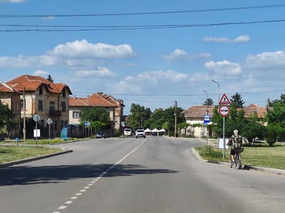 The image depicts a view of a street in Krushovitsa, Bulgaria, on a sunny day with scattered clouds. A paved road curves through a village setting, lined with traditional houses featuring red tile roofs. Several cars are visible, including a black car in the distance, a white van, and a white SUV. A cyclist, wearing a camouflage shirt and dark shorts, is riding away from the camera on the right side of the road. Traffic signs are present, including a pedestrian crossing sign and a no-passing sign. A blue bus stop shelter is situated on the left side of the road, near a light blue fire hydrant. Trees and greenery are abundant throughout the scene, contributing to the rural atmosphere.