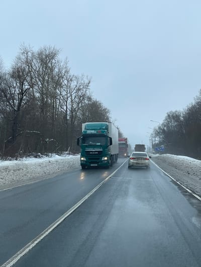 The image depicts a winter scene on a wet, two-lane highway in the city of Podvyaznovo, Russia, under an overcast sky. A green MAN semi-trailer truck, featuring "ТВЭЛ | TVEL" and "ЛОМОВСКИЙ | LOMOWSKY" branding, with Russian license plate "P6 929", drives in the left lane towards the viewer with its headlights illuminated. Ahead in the right lane, a light-colored sedan proceeds away from the viewer, its rear license plate obscured by debris. Following the sedan is a red semi-trailer truck, also moving away, with another dark truck visible further in the background. The road surface is dark and reflective from moisture, separated by a solid white line. Accumulations of snow line both shoulders of the highway. Bare deciduous trees form dense wooded areas flanking both sides of the road. Utility poles with overhead lines and streetlights are visible along the right side of the highway, along with a partially visible blue road sign in the distance. No individuals are visible outside of the vehicles.