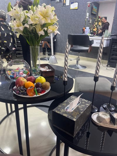 This image depicts the interior of a hair salon located in Talin, Armenia.
In the immediate foreground, two black circular tables with black metal legs are positioned. The table on the left holds a tall clear glass vase containing a large bouquet of artificial white lilies with yellow accents. Next to the vase is a clear glass fishbowl filled with various colorful wrapped candies. An oval white plate on this table displays an arrangement of fresh fruits including red apples, oranges, plums, kiwi, grapes, and a partially eaten persimmon, alongside which rests a small green chili pepper. A small brown woven basket containing wrapped chocolates or snacks is also on this table.
The table on the right, slightly lower, features three silver, twisted, tapered candles in black metal holders, with two fully visible. A rectangular tissue box with a dark, patterned design and a white tissue protruding is also on this table. Adjacent to it, a light gray hexagonal tray holds two small hexagonal candles or holders and a dark card.
In the midground, a black, height-adjustable salon chair with a chrome base stands empty. Beyond this, the walls are covered with dark gray textured wallpaper. On the far left, the back of an individual, presumably a client, is visible, seated in a salon chair and draped with a dark cape adorned with a feather pattern. Their gray hair is partially visible. Further back, two light gray dome-style hair dryers are present on the floor.
To the right, a large wall mirror reflects a male individual, likely a stylist, wearing a dark brown long-sleeved shirt. He is standing at a workstation, facing away from the camera. Also reflected in the mirror is a decorated Christmas tree with lights and ornaments, positioned behind the stylist. Certificates or framed pictures are visible on the wall above the empty salon chair. A light gray upholstered cube or ottoman is situated between the tables and the empty salon chair.