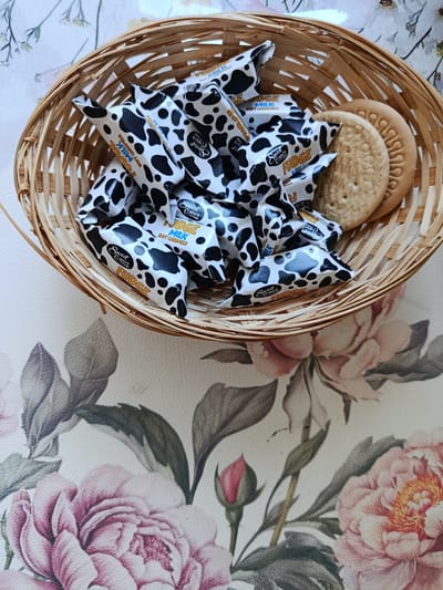 A woven basket filled with individually wrapped candies with a cow print design and two round biscuits rests on a white surface with a floral pattern. The candies are wrapped in white paper with black cow spots and feature text such as "Sweet Corner FUDGE MILK SOFT CARAMELS". One biscuit is partially visible to the right of the candies, and another is placed behind it. The floral pattern on the surface features pink roses and green leaves. The image is a top-down view, and no specific location in Pardubice, Czechia can be identified.