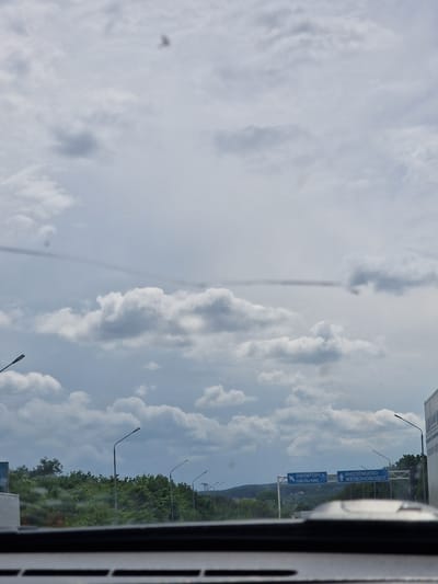 This is a photograph taken from inside a vehicle, looking out at a cloudy sky and a landscape with trees and signs. The signs indicate directions to "ПЯТИГОРСК НАЛЬЧИК" (Pyatigorsk, Nalchik) and "ИНОЗЕМЦЕВО ЖЕЛЕЗНОВОДСК" (Inozemtsevo, Zheleznodorozhny). This indicates the location is near the crossroads for these cities, consistent with the area around Змейка, Russia, which is a rural locality within the city of Zheleznodorozhny. The scene is a roadside view, with streetlights visible along the road and a dense green tree line in the middle ground. The sky is predominantly filled with white and grey clouds. There are no people or overt actions depicted in the image.