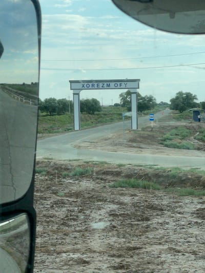 The image shows a road in a rural area. The camera is positioned within a vehicle, likely a car, as a side mirror is visible in the foreground. The landscape is dry and dusty, with sparse vegetation and some trees in the distance. A road stretches into the distance, with a bus stop sign visible along its side. There is also a sign with text that reads "XOREZM OFY". The sky is cloudy and appears to be daytime. The overall impression is of a journey through a semi-arid region.