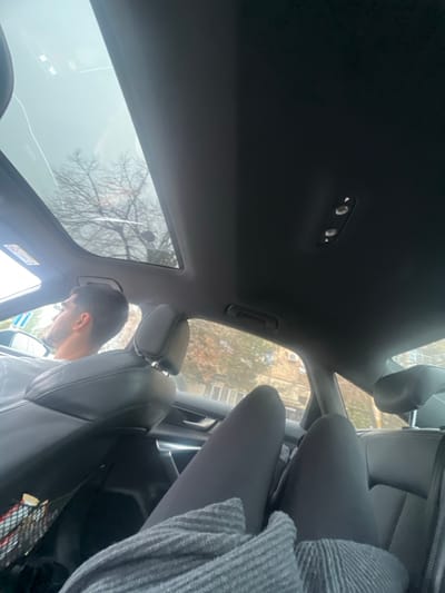 The image displays the interior of a vehicle, observed from a rear passenger's perspective, located within Stara Pazova Municipality, Serbia. A male individual, with short dark hair and a beard, occupies the driver's seat, visible from the chest upwards, wearing a light-colored top. His head is oriented slightly to the left. In the foreground, the legs of a second individual are visible, extended, clothed in dark leggings and partially covered by a grey, ribbed textile.
The vehicle's interior features dark leather seating, a dark headliner, and a panoramic glass sunroof through which a clear view of the sky and tree branches is visible; a linear anomaly, possibly a crack or reflection, crosses the sunroof. An overhead console with two circular lights/buttons is present. Through the vehicle windows, an outdoor environment is discernible, including bare deciduous trees and multi-story buildings with rectangular windows. A portion of a road sign is visible through the driver's side window. The back of the driver's seat contains a mesh pocket with indistinct contents. The driver appears to be operating the vehicle, while the rear passenger's posture suggests a seated, relaxed position.