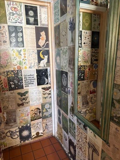 The image shows an interior ceiling and upper wall space covered in a collage of printed images. The collage appears to be composed of vintage illustrations and text, with themes relating to the moon, nature, cats, and occult symbols. The collage is organized in a grid-like fashion, with individual images cut and placed to cover the surfaces. The ceiling area is framed by a greenish-painted trim. Some areas near the corner of the upper wall suggest a connection to an adjacent area with plain tiles. The location is difficult to ascertain with certainty, but given the information provided, the image could be from a place within West Oxfordshire, UK. No people are depicted.