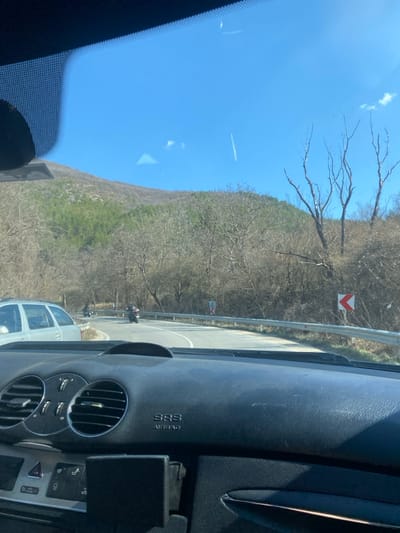 The image captures a scene from inside a vehicle, looking out through the windshield onto a winding mountain road in Konyavo, Bulgaria, on a clear, sunny day. The foreground shows the vehicle's dark gray dashboard, featuring circular air vents on the left, control buttons, an "SRS AIRBAG" label on the passenger side, and a black mounting device on the lower right. Reflections are present on the windshield.
The asphalt road curves to the right, with a metal guardrail lining its right edge. Behind the guardrail, a red and white chevron sign indicates the sharp right turn. Three individuals on motorcycles are visible on the road ahead, navigating the curve. The most prominent motorcyclist is in the center, wearing dark gear, with two other motorcyclists further behind. A silver-colored station wagon is partially visible on the left side of the road, appearing to be stationary or moving slowly.
The background is dominated by a range of hills or mountains covered predominantly with bare deciduous trees, suggesting late winter or early spring. Patches of green evergreen trees are visible on the upper slopes to the left. The sky is clear and blue, with a faint white contrail from an aircraft visible in the upper right quadrant. Several prominent bare trees with distinct upward-reaching branches are positioned on the right side of the scene, behind the guardrail.