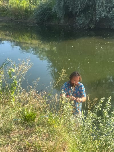 A middle-aged man with shoulder-length brown hair and a beard is standing at the edge of a body of water. He is wearing a short-sleeved, brightly colored Hawaiian shirt and light-colored shorts. The man is holding something in his hands and appears to be examining it closely. The water is dark green and reflects the surrounding vegetation and sky. The foreground is filled with tall, dry grass and green bushes. In the background, there are more trees and lush green foliage along the bank of the water. The image is taken outdoors during daylight.