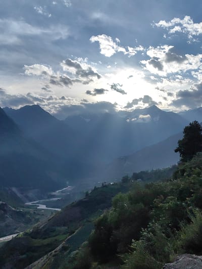 The image depicts a dramatic mountain landscape under a cloudy sky, with sunbeams breaking through the clouds and illuminating the scene. In the foreground, there is lush green vegetation and a dark tree on the right side. The midground features rolling hills with patches of vegetation and scattered structures, suggesting a rural or village setting. A winding river flows through the valley in the midground. The background is dominated by majestic, layered mountains fading into a misty blue haze, with some peaks possibly snow-capped. The overall impression is one of natural grandeur and a remote, possibly high-altitude location. Based on the provided information, the scene is located in Tiling, India.
