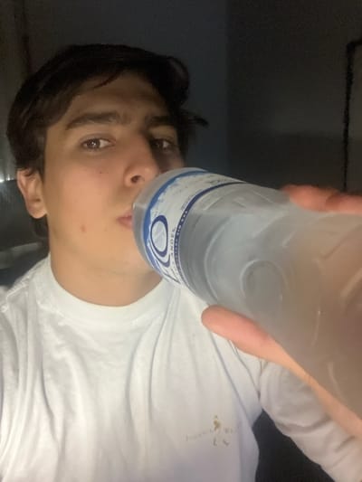 A person is drinking from a clear plastic water bottle. The person has dark hair and light skin, wearing a white t-shirt with a logo partially visible, "Johnnie Walker." The background is a dark, indistinct space. The bottle has a blue and white label with the word "ANDES" visible. The location, Gerli, Argentina, is not directly discernible from the visual elements in the photo.