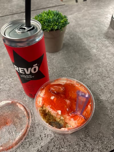 The image depicts a tabletop scene. On a grey textured surface, there is a red can of "REVO" brand alcohol energy drink with a black straw inserted. Adjacent to the can is a small potted green plant. Next to the plant is a clear plastic container filled with a dessert, consisting of a reddish jelly-like topping and a granular base. A purple plastic spoon is partially embedded in the dessert. A matching lid for the container is placed to the right. There are no people visible in the image. The setting is unknown, but the absence of any specific landmarks or indicators in the image makes the determination of a location in Satovcha, Bulgaria impossible.