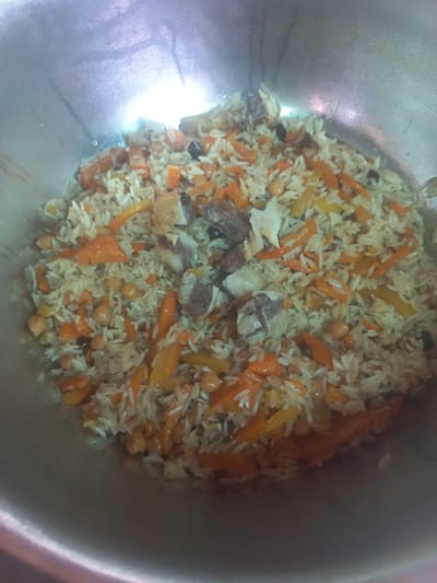 A close-up, top-down view shows a metal bowl filled with plov, a traditional Uzbek rice dish. The plov is a mixture of white rice, julienned carrots, chickpeas, and chunks of meat. The meat appears to be lamb or beef, with some pieces showing bone. The carrots are bright orange and interspersed throughout the dish. The chickpeas are a lighter tan color. The overall color palette of the plov is warm and earthy. The metal bowl is large and appears to be a cooking pot or serving dish. The surface of the bowl is somewhat reflective, indicating it is likely made of stainless steel or aluminum. The lighting in the image is bright and overhead, creating some glare on the surface of the bowl. There are no people visible in the image. The scene is presented in a way that focuses solely on the food itself. The location in Yorkatay, Uzbekistan, is implied by the dish being depicted.