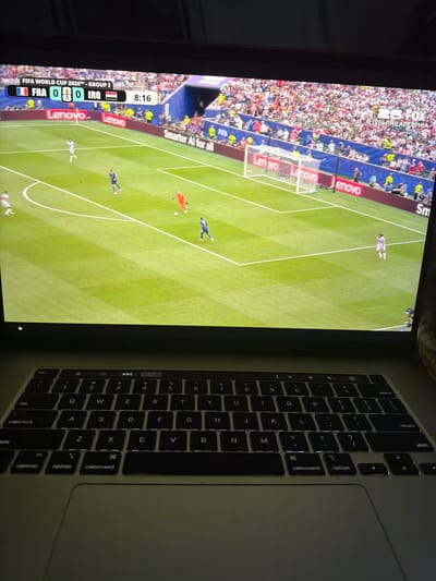 The image shows a close-up view of a laptop screen displaying a soccer match. The screen shows a game between FRA (France) and IRQ (Iraq) with the score at 0-0 in the 8th minute. The match is identified as FIFA WORLD CUP 2026™, GROUP I. The game is being broadcast on FOX, with "LIVE PHILADELPHIA" visible in the top right corner of the screen. The soccer field is green with white lines, and players in blue and white uniforms are visible on the field. There are also advertisements for Lenovo and Smarter AI for all around the stadium. The laptop keyboard is in the foreground, with the keys illuminated. The overall setting appears to be indoors, possibly a home or office, given the presence of the laptop. The city of Fulshear, United States of America, is not depicted or referenced in the image.