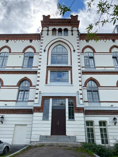 The image shows the exterior of a building with a distinct architectural style, featuring white walls accented with red brickwork. The building has a multi-story facade with numerous windows, some of which are arched or circular. Decorative brick trim and dentil molding are visible along the roofline and between stories. A prominent, large arched window is a focal point on one section of the building. The building's entrance appears to be a dark brown door with a metal handle. The setting seems to be an outdoor urban or semi-urban environment. There are elements of nature, with tree branches and leaves partially obscuring the upper left portion of the image, and greenery visible along the right side. The sky above is partly cloudy, suggesting daytime. The overall impression is of a historical or classically designed building. The image is rotated 90 degrees counter-clockwise.