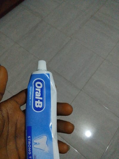 The image depicts a human hand holding a tube of Oral-B toothpaste against a tiled floor background in the city of Abatete, Nigeria. The hand, with a dark skin tone, is positioned centrally, gripping the blue and white toothpaste tube with its thumb and fingers. The visible text on the tube includes "Oral-B", "TOOTHPASTE", and "STRONG TEETH", accompanied by a graphic of a white tooth. The toothpaste tube has a white, ridged screw-on cap. The background consists of light gray or beige square floor tiles with visible grout lines. A distinct bright reflection is visible on the lower right portion of the tiled floor, indicating an overhead light source.