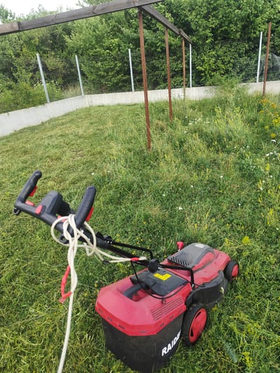 The image depicts a red electric lawnmower positioned on a grassy area, likely in Prolesha, Bulgaria. The lawnmower is the central subject, with its handle and cord visible in the foreground. The grass appears to be uncut in some areas and recently mowed in others, suggesting recent activity. In the background, a wire fence with concrete supports and a wooden structure are visible, bordering a dense area of trees and foliage. The overall scene suggests an outdoor residential or garden setting. There are no people or animals present in the image.