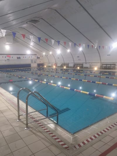 This media file shows an indoor swimming pool. The scene is set in a large, well-lit hall with a high, arched ceiling. The pool itself is a standard competition-style pool with lanes marked by floating ropes in red, white, and blue. The water is a vibrant blue, and reflections of the overhead lights create a shimmering effect.

Along the sides of the pool, there are tiled surfaces and metal railings leading into the water. Benches are visible along the far wall, suggesting spectator or rest areas. String lights and a digital clock displaying "15:19" are mounted on the wall above the pool, indicating the time. Bunting in red, white, and blue is strung across the ceiling, adding a festive touch. The overall impression is that of a public or training facility, likely operational. The location cues, such as the text "Қосшағыл ауылдық округі, Kazakhstan," suggest the specific geographic context. The weather is not discernible as the image is of an indoor setting. There are no people visible in the frame, so no emotions can be inferred from their presence.