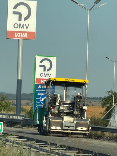 In Kaspichan, Bulgaria, an asphalt paving machine, branded "VÖGELE," is captured on a sunny day, operating on a road surface. The machine is grey and black with a yellow canopy. To the left of the paving machine, two OMV petrol station signs are visible. The larger sign, positioned further left, is white with a blue "OMV" logo and a red "VIVA" band. The smaller sign, closer to the paving machine, has a green border and a white background with a blue "OMV" logo. In the background, a tall street lamp with three lights stands to the right, and another is partially visible to the left. The surrounding environment consists of dry grass, a guardrail, and distant fields.