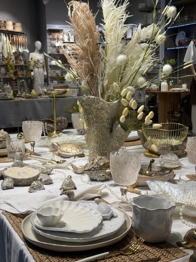 The image depicts a highly stylized tablescape, likely staged for a decorative or retail display. The location is indicated as Novoalexandrovka, Russia, but this does not appear visually substantiated within the image itself.
The primary focus is a table setting with a light-colored tablecloth. A centerpiece features a vase containing feathery plumes and dried floral elements. The table is adorned with various decorative items, including stylized turtles, faux pearls, patterned glassware, and what appears to be decorative wood slices. The place settings include white plates with shell-like designs and metallic flatware. A woven runner sits beneath part of the arrangement.
Reflections in the background suggest the presence of a retail environment, potentially a home goods store or showroom, with shelves displaying additional items. A mannequin is also visible in the reflected background. There are no people directly visible in the main focal area of the image.