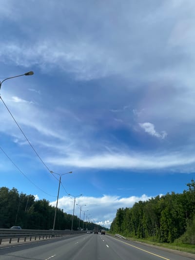 The image shows a view of a highway, likely captured from a moving vehicle. The scene includes a clear blue sky with wispy clouds, with a distinct separation of cloud formations. A row of street lights lines the side of the road, interspersed with trees on the embankment. The highway is divided by a metal barrier. Cars are visible on the roadway. Given the location is near Serp I Molot, Russia, it is possible that this scene depicts a highway in that region.