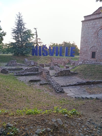 A large blue sign spelling "NIŠVILLE" is positioned behind ancient stone ruins in the city of Niš, Serbia, specifically within the Crveni Krst municipality. To the right of the ruins, a portion of a building constructed from stone and brick is visible, featuring a arched window with a decorative lattice. The foreground of the image is a grassy area with scattered stones and fallen leaves. In the background, a tall coniferous tree stands behind the sign, and more trees are visible further in the distance, against a pale sky. The overall scene depicts an archaeological site with a modern landmark.