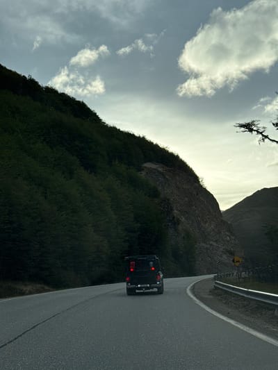 A dark-colored utility vehicle, likely an SUV, travels away from the viewer on a two-lane asphalt road that curves gently to the right. Its two red taillights are illuminated. Two red containers, possibly jerry cans, are mounted on the vehicle's rear, which also displays a tow hitch. The road features a solid white line on its right edge. To the left of the road, a steep, densely forested hillside rises, transitioning to a rocky slope higher up. To the right of the road, a metal guardrail follows the curve, and a yellow diamond-shaped road sign with a black curved arrow is visible. The sky above is partly cloudy, displaying light grey and blue tones, suggesting either dawn or dusk. Distant mountainous terrain is visible in the background. The scene is located in the city of Lago Escondido, Argentina.