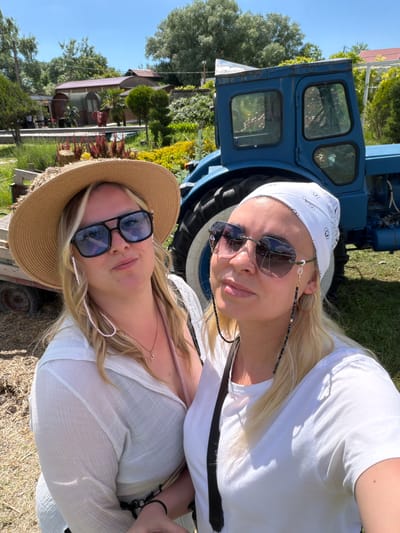This outdoor photograph, taken in Gay-Kodzor, Russia, features two women posing in a sunny, rural setting.
On the left, a woman with blonde hair wears a white bandana adorned with small, repeating black motifs. She has on dark, rectangular sunglasses connected by a black and white beaded chain, a large gold hoop earring, and a white short-sleeved t-shirt. She looks towards the camera with a neutral expression.
To her right, another woman with blonde hair is wearing a wide-brimmed straw hat and dark, square-framed sunglasses, which are connected by a light pink beaded chain. She is dressed in a flowing, white, sheer-textured top or dress and also looks towards the camera.
In the left background, partially obscured by the women, is a blue tractor or similar agricultural vehicle, revealing its side and part of its cabin with multiple window panes. The background is filled with lush greenery, including various trees, shrubs (some evergreen, some with red leaves), and grassy ground. Further back, a building with a reddish roof, possibly a house or shed, is visible amongst the foliage. The bright blue sky and strong sunlight, casting clear shadows, suggest a midday or early afternoon setting. The overall scene depicts a relaxed outdoor moment. No discernible text is visible, though the bandana features decorative patterns.