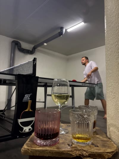In a room with concrete walls and a suspended ceiling featuring industrial lighting, a man is actively engaged in a game of table tennis. He is wearing a light-colored t-shirt and shorts, positioned on the right side of the image, facing away from the viewer and towards the table. The table tennis table, with its net, occupies the central and left portions of the frame. In the foreground, three glasses are arranged on a rough-hewn wooden surface. The leftmost glass contains a dark red liquid, likely wine. Next to it, a stemmed wine glass holds a pale yellow liquid, presumably white wine. The rightmost glass contains a light brown liquid, possibly an alcoholic beverage. The background shows piping along the ceiling. The overall scene suggests a casual indoor recreation activity.