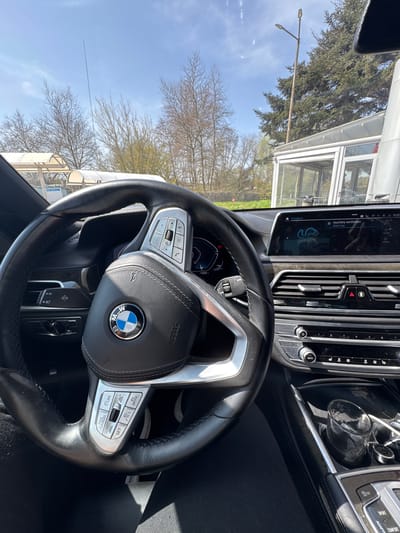 The image depicts the interior of a BMW, specifically the steering wheel and dashboard. The car is equipped with advanced features, as evidenced by the multiple buttons and controls on the steering wheel and center console. The dashboard screen displays information about auxiliary ventilation and temperature settings. Outside the car window, there are trees, a street lamp, and a building with glass windows, possibly a commercial establishment. Although the image was supposedly taken in Staro Oriahovo, Bulgaria, there's nothing in the image that explicitly confirms that.