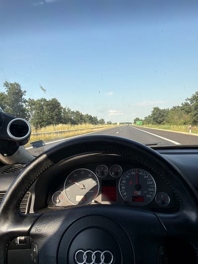 The image depicts a driver's perspective from within an Audi S4 traveling on a highway in the city of Svoboda, Bulgaria, under a clear daytime sky. The steering wheel, featuring a black leather wrap and the Audi rings emblem, is visible in the lower portion of the frame. The instrument cluster behind the steering wheel displays a speedometer indicating approximately 112-115 km/h, an odometer reading of 299308, and an S4 badge. Additional gauges for RPMs, water temperature, fuel level, and voltage are present. An aftermarket circular gauge, likely a boost gauge, is mounted on the left side of the dashboard. The view through the windshield reveals a clean asphalt highway with at least two lanes in the direction of travel, bordered by a grassy median and roadside foliage consisting of trees and yellow wild plants. White and red reflective marker posts line the right side of the road. A green overhead road sign is faintly visible in the distance. The windshield exhibits numerous small specks of dirt and a larger, elongated greenish mark on the upper left. No individuals are explicitly visible.