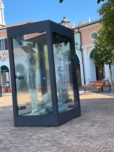 This image shows a storefront display at the Noventa di Piave Outlet in Noventa di Piave, Italy. The display is a large glass box with a dark frame, tilted slightly. Inside, a mannequin is dressed in a bikini top and a long, patterned skirt featuring wavy stripes of blue, white, and orange. The background inside the display is a chevron pattern. The name "MISSONI" is visible on the lower right side of the display. Outside the display, the ground is paved with red bricks laid in a herringbone pattern. In the background, there are buildings with pastel-colored facades and architectural details. Two wooden benches are positioned in front of one of the buildings, and a tree with green foliage stands to the right of the display. A sign for "JO MALONE LONDON" is visible on a building to the left, with a few people walking in that area. The sky is clear and blue.