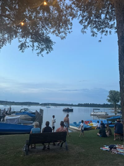 At a lakefront in Rimsting, Germany, an outdoor gathering unfolds under a twilight sky. The scene is dominated by a calm body of water dotted with various boats, including sailboats, a motorboat, and paddle boats. Along the shore, a row of docked boats, some covered with blue tarpaulins, is visible.
Several groups of people are scattered across the grassy area. In the foreground, three individuals are seated on a wooden bench, facing away from the camera. To their right, a group of people are sitting on blankets on the grass, and a boy is walking on the lawn. Further down the shore, a woman walks towards the water, and a group of people are gathered near the paddle boats. In the distance, a line of trees and buildings forms the shoreline. The overall atmosphere suggests a relaxed evening, possibly a barbecue or social event by the water.